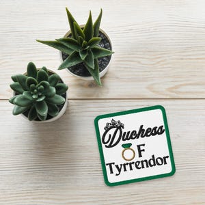 Pu&ograve; includere: Toppa quadrata con bordo verde e le parole "Duchess of Tyrrendor" in corsivo nero. &Egrave; presente un anello d'oro con una gemma verde. Nell'immagine sono presenti anche due piccole piante grasse in vaso.