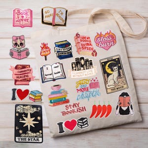 May include: A beige canvas tote bag decorated with numerous embroidered patches featuring book-themed illustrations, hearts, chili peppers, and tarot card designs. The patches include text such as "Book Lover," "Slow Burn," "Certified Bookworm," "I Cannot Live Without Books," "Just One More Chapter," "Stay Bookish," and "The Star."