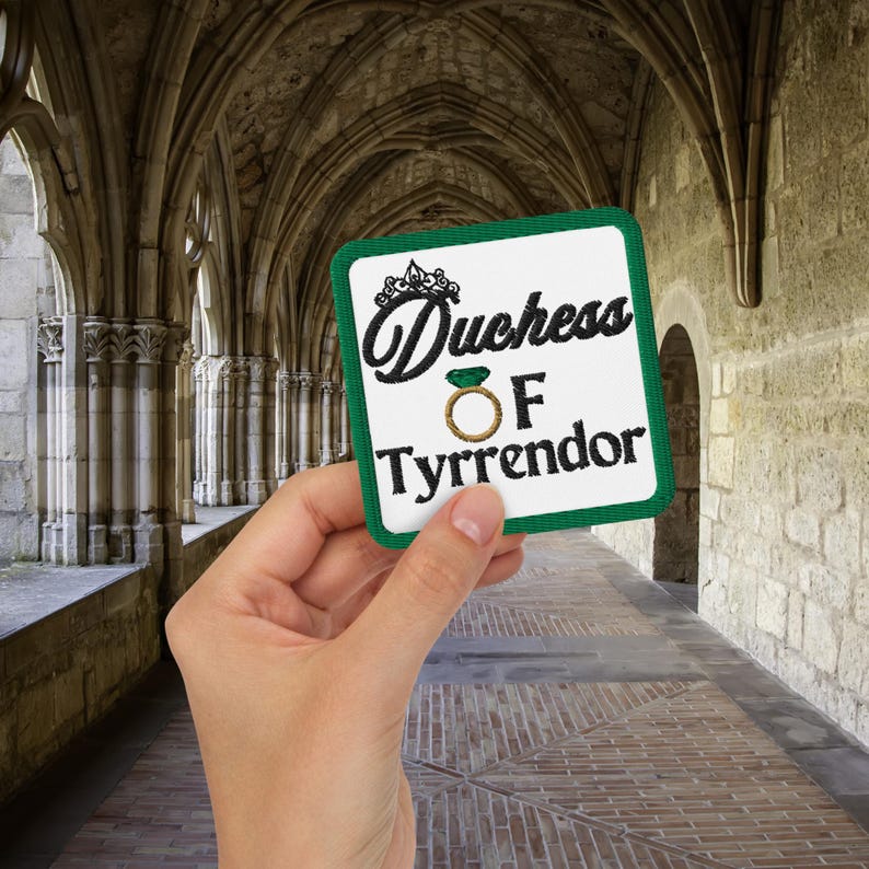 Pu&ograve; includere: Una toppa quadrata con bordo verde e le parole "Duchess of Tyrrendor" in corsivo nero. La toppa presenta una corona e un disegno ad anello. La toppa &egrave; tenuta in mano su uno sfondo di arcate in pietra.