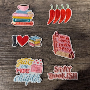 May include: A collection of six embroidered patches featuring a stack of books with a cup, four red chili peppers, a heart with books, a book labeled I LOVE MY BOOKS EXTRA SPICY, a book with text JUST ONE MORE CHAPTER, and text STAY BOOKISH.