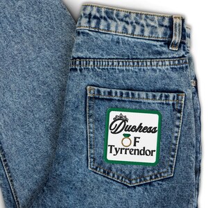 Pu&ograve; includere: Jeans blu chiaro con una toppa quadrata sulla tasca posteriore. La toppa ha uno sfondo bianco con le parole "Duchess OF Tyrrendor" in nero e una grafica ad anello dorato.