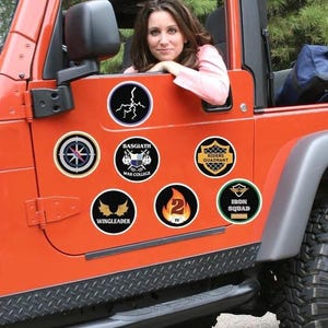 May include: A bright orange Jeep with a variety of round stickers on the side. The stickers feature different designs, including a compass, a military insignia, a winged creature, a number "2" with flames, and a green sticker with the words "Iron Squad".