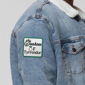 Pu&ograve; includere: Giacca di jeans blu chiaro con colletto in montone sintetico bianco. Una toppa verde e bianca sulla giacca recita "Duchess of Tyrrendor". La giacca ha due tasche frontali e bottoni scuri.