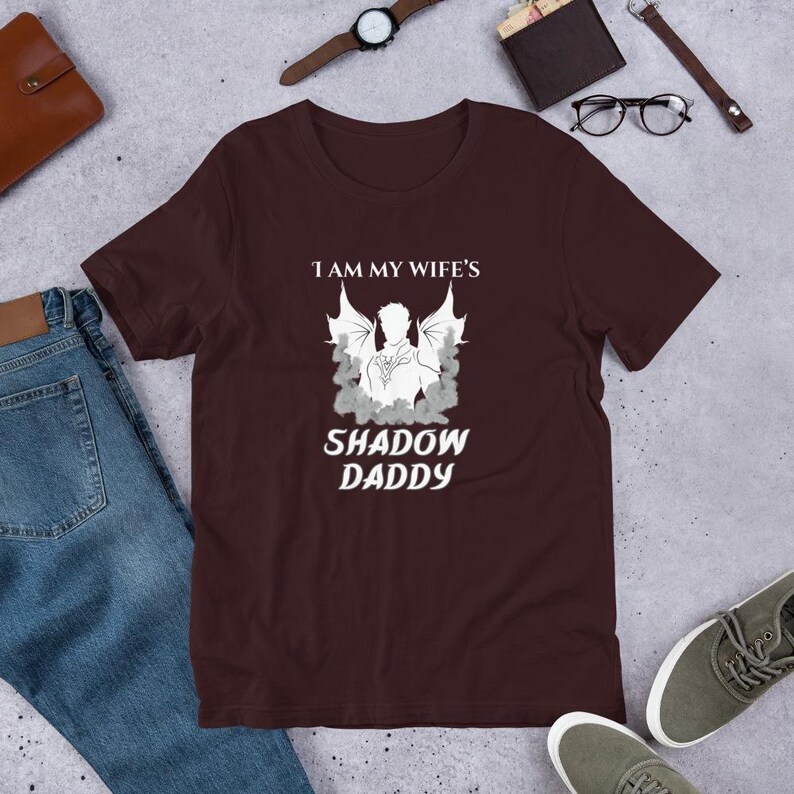 Pu&ograve; includere: Una maglietta bordeaux con la scritta bianca "I AM MY WIFE'S SHADOW DADDY". La grafica presenta la silhouette di una figura con le ali e uno sfondo a forma di nuvola.