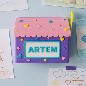 Könnte beinhalten: Ein lila-rosa Briefkasten mit dem Namen "ARTEM" in weißen Buchstaben. Der Briefkasten ist mit Herz- und Sternformen verziert. Zeichnungen eines Schneemanns, eines Hauses und eines Fisches sind an der hellblauen Wand befestigt.