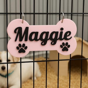 May include: A pink bone-shaped pet tag with the name "Maggie" in black script and two black paw prints. The tag hangs in a black wire cage, with a small dog house and a puppy in the background.