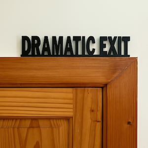 Può includere: Un cartello nero con il testo DRAMATIC EXIT in un carattere grassetto senza grazie. Il cartello è posizionato orizzontalmente sopra una cornice di porta in legno con una finitura marrone chiaro naturale e venature visibili su una parete bianca.