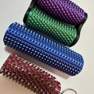 May include: Four massage rollers in shades of purple, green, blue, and red. Each roller features a textured surface with a repeating pattern. Designed for muscle relief, one roller includes a keyring.