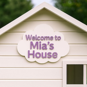 Op de afbeelding: Een crèmekleurig speelhuisje met een wit dak en gevelbekleding. Een wolk-vormig bord met de woorden "Welcome to Mia's House" in paarse letters is bevestigd aan de voorkant van het speelhuisje.