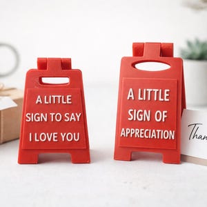 May include: Two red, A-frame miniature signs with white raised lettering. The left sign reads, A LITTLE SIGN TO SAY I LOVE YOU. The right sign reads, A LITTLE SIGN OF APPRECIATION. Both signs feature a handle cutout at the top and a hinged design.