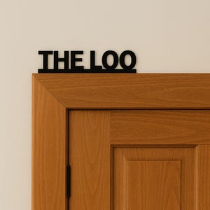 May include: A black metal sign reading "THE LOO" above a wooden door. The sign is in a bold, sans-serif font. The door is made of light brown wood with a visible grain. A simple, stylish bathroom sign.