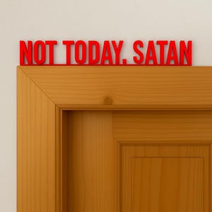 Pode incluir: Placa de madeira vermelha com a frase "NOT TODAY, SATAN" montada acima de uma porta de madeira. A placa está em uma fonte em negrito, sem serifa. A porta é de cor marrom claro.