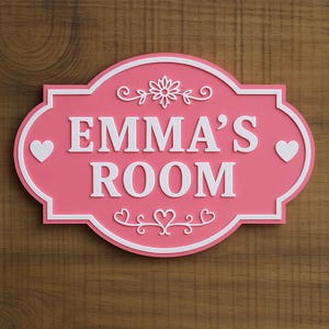May include: A pink oval-shaped wall sign with a white border and white text that reads EMMA'S ROOM. The sign features a white floral flourish above the text, a white heart on each side, and a white decorative scroll design below the text.