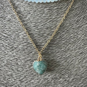 May include: A gold chain necklace with a green heart-shaped pendant. The necklace is layered over a white beaded necklace.