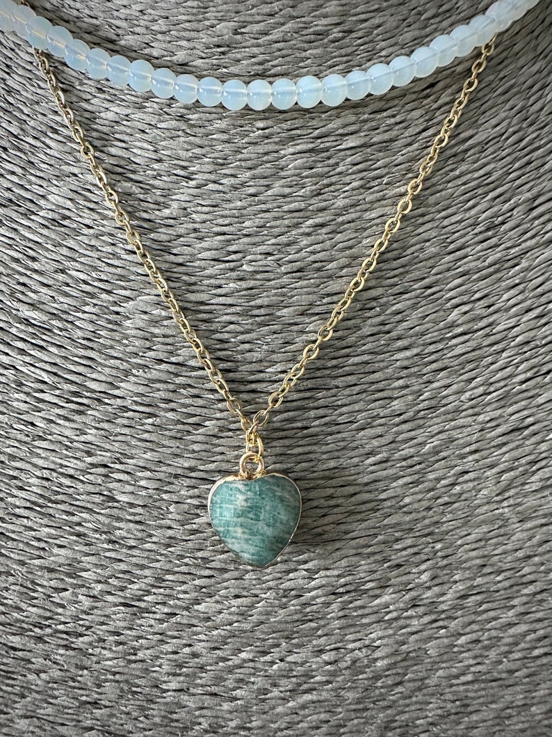 May include: A gold chain necklace with a green heart-shaped pendant. The necklace is layered over a white beaded necklace.