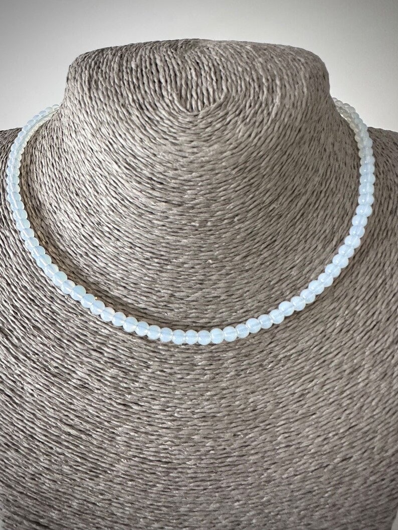 May include: A delicate white beaded necklace on a mannequin bust. The necklace is made of small, round beads that are strung together on a thin cord.