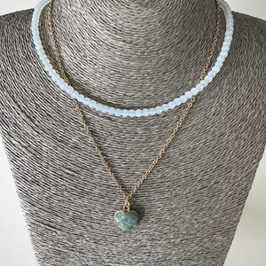 May include: A layered necklace with a delicate gold chain and a heart-shaped pendant. The pendant is made of a green stone with a marbled pattern. The top layer of the necklace is made of white beads.