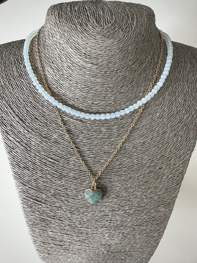 May include: A layered necklace with a delicate gold chain and a heart-shaped pendant. The pendant is made of a green stone with a marbled pattern. The top layer of the necklace is made of white beads.