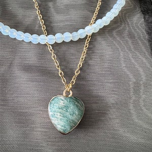 May include: A gold chain necklace with a green heart-shaped pendant. The necklace is layered over a white beaded necklace.