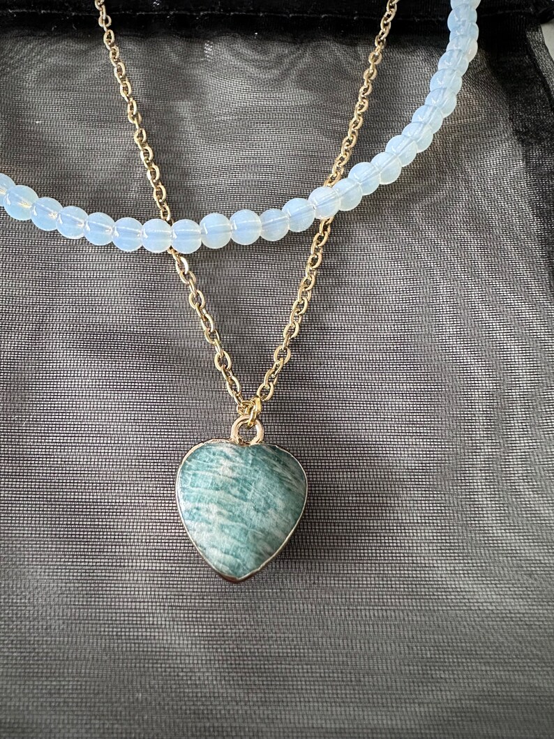 May include: A gold chain necklace with a green heart-shaped pendant. The necklace is layered over a white beaded necklace.