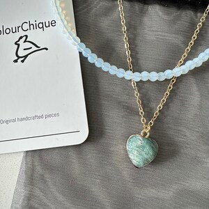 May include: A delicate layered necklace with a heart-shaped green gemstone pendant. The necklace is displayed on a black mesh bag with a white card that reads "ColourChique" and "Original handcrafted pieces".