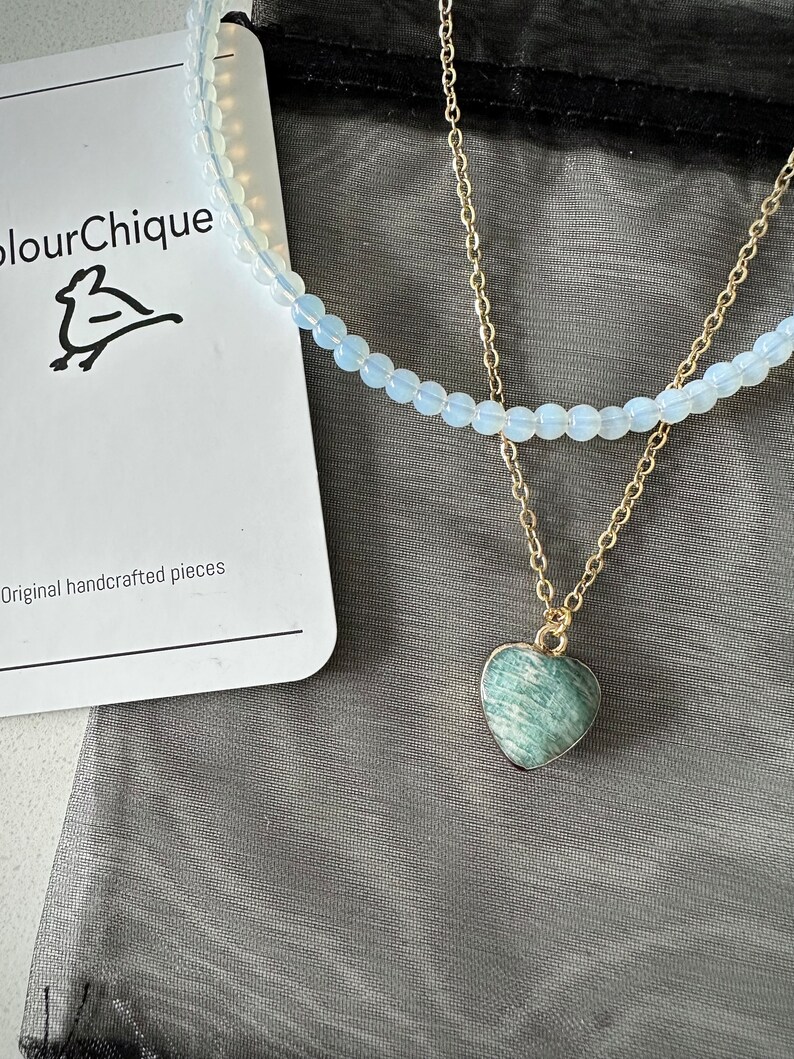 May include: A delicate layered necklace with a heart-shaped green gemstone pendant. The necklace is displayed on a black mesh bag with a white card that reads "ColourChique" and "Original handcrafted pieces".