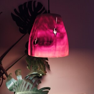 Peut inclure: Un luminaire suspendu avec un abat-jour marbré rose et violet. L'abat-jour a une forme organique unique avec des veines de bois et des nœuds naturels visibles. La lumière est suspendue à un câble noir, dégageant une lueur chaleureuse.