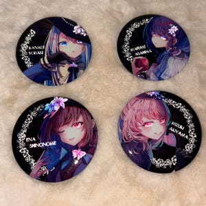 Puede incluir: Cuatro botones redondos negros y plateados con imágenes de personajes de anime. Cada botón presenta un personaje diferente con un diseño y un nombre únicos. Los personajes son Kanade Yosak, Maruyo Asamina, Ena Shinonome y Mizuki Akiyama.