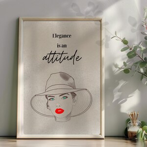 Könnte beinhalten: Gerahmter Kunstdruck mit einer Skizze einer Frau mit breitkrempigem Hut, roten Lippen und türkisfarbenen Augen. Der Text "Elegance is an attitude" steht in stilvoller Schrift über der Illustration.