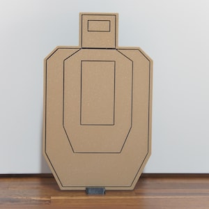 May include: A tan cardboard shooting target with a human-like silhouette shape. The target features black printed outlines of a head box and a central torso zone, designed for marksmanship practice. The material has a textured, matte finish.