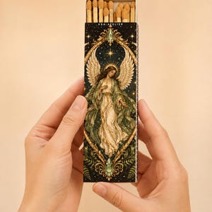 May include: A rectangular matchbox featuring a dark background with a central illustration of an angel with large wings, surrounded by a gold ornate frame and green accents. The text KEA ATELIER is printed at the top. The box is filled with wooden matches.
