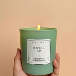 May include: A lit green candle in a cylindrical glass jar. The candle has a white label with the text "KEA ATELIER", "SUNSHINE KISS", and other details. The candle is held by a person.