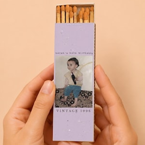 May include: A rectangular matchbox with a light purple speckled cover featuring a photograph of a child in a cream vest and blue pants. The text reads Sarah's 30th birthday and VINTAGE 1996. The box is filled with wooden matches with brown tips.