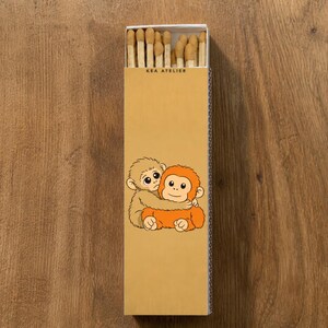K&ouml;nnte beinhalten: Eine rechteckige Streichholzschachtel mit einer Cartoon-Illustration von zwei sich umarmenden Affen. Die Schachtel ist beige mit dem Text "KEA ATELIER" oben aufgedruckt. Die Streichholzk&ouml;pfe sind in der Schachtel sichtbar.