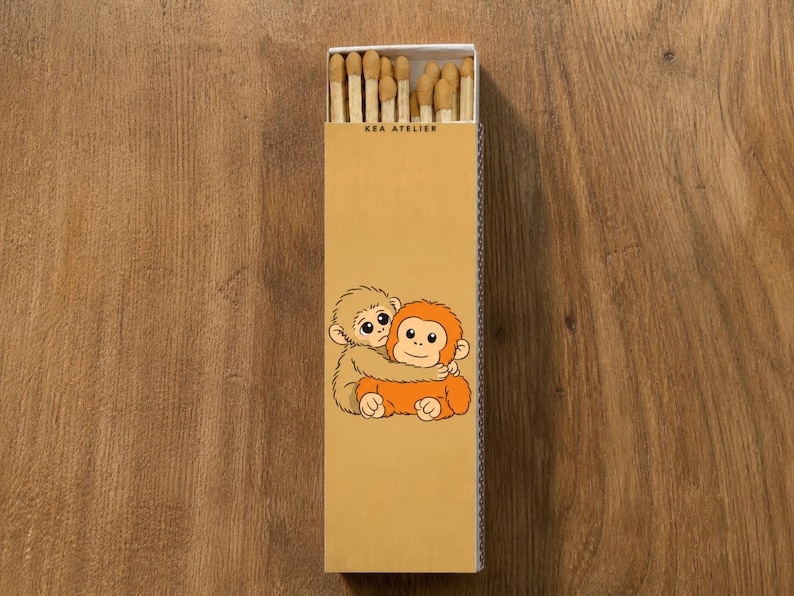 K&ouml;nnte beinhalten: Eine rechteckige Streichholzschachtel mit einer Cartoon-Illustration von zwei sich umarmenden Affen. Die Schachtel ist beige mit dem Text "KEA ATELIER" oben aufgedruckt. Die Streichholzk&ouml;pfe sind in der Schachtel sichtbar.
