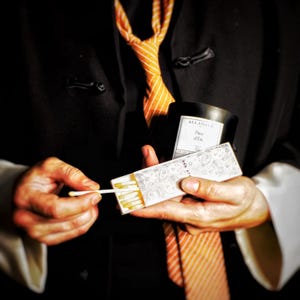 May include: A person holding a matchbox and a black candle. The matchbox is open, revealing several matches. The candle has a label that reads "Fleur d'&Eacute;t&eacute;". The person is wearing a black jacket and an orange striped tie.