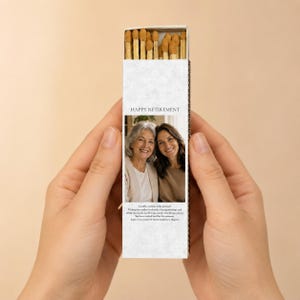 May include: A rectangular matchbox with a white textured cover featuring the text HAPPY RETIREMENT and a photograph of two smiling individuals. The box is filled with wooden matches that have tan tips, visible at the top of the open sliding tray.