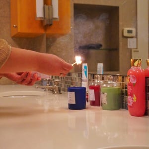 May include: A lit match ignites a blue candle, with other candles and bath products in the background. The scene is set in a bathroom with a neutral color palette. The candle is cylindrical and has a white label.