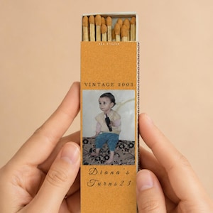 May include: A matchbox with a vintage design. The box is tan with the text "VINTAGE 2003" and a photo of a child. The text "Diana's Turns 23" is below the photo. The match heads are visible.
