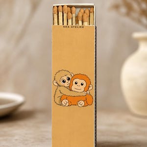 K&ouml;nnte beinhalten: Eine rechteckige Streichholzschachtel mit dem Text "KEA ATELIER" oben. Die Schachtel zeigt eine Cartoon-Illustration von zwei sich umarmenden Affen in Braun- und Oranget&ouml;nen. Die Schachtel ist mit Holzstreichh&ouml;lzern gef&uuml;llt.