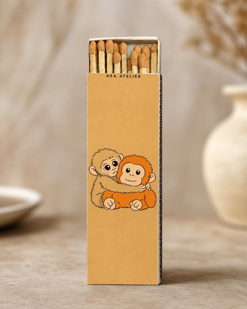K&ouml;nnte beinhalten: Eine rechteckige Streichholzschachtel mit dem Text "KEA ATELIER" oben. Die Schachtel zeigt eine Cartoon-Illustration von zwei sich umarmenden Affen in Braun- und Oranget&ouml;nen. Die Schachtel ist mit Holzstreichh&ouml;lzern gef&uuml;llt.