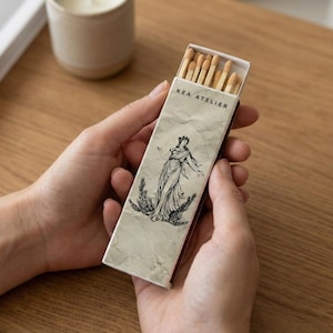 May include: A rectangular matchbox held in two hands, featuring the text "KEA ATELIER" above a black and white illustration of a woman in a flowing gown. The matchbox is open, revealing the matchsticks.