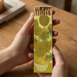May include: A matchbox featuring a print of Van Gogh's "Sunflowers" painting. The box is held by hands and contains several wooden matches with brown tips. The artwork is predominantly yellow and green, with a white vase.