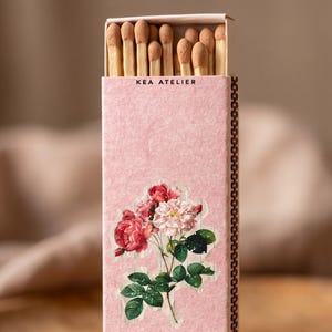 May include: A rectangular matchbox with a textured light pink cover featuring a floral illustration of red and white roses with green leaves. The text KEA ATELIER is printed in black near the top. The box is partially open, revealing wooden matches.