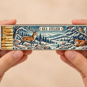 May include: A rectangular matchbox featuring a winter forest illustration with a deer, a fox, and an owl. The box is labeled KEA ATELIER and contains wooden matches with tan tips. The artwork depicts snow-covered pine trees and mountains.