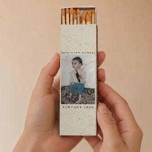 May include: A rectangular matchbox with a light beige speckled finish containing wooden matches with tan tips. The front features a photograph of a child, the text Sarah's 30th birthday, and the text VINTAGE 1996 printed in a black serif font.