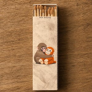 May include: A matchbox with the text "KEA ATELIER" features a cartoon illustration of two hugging monkeys. The match heads are visible at the top. The box is on a wooden surface.