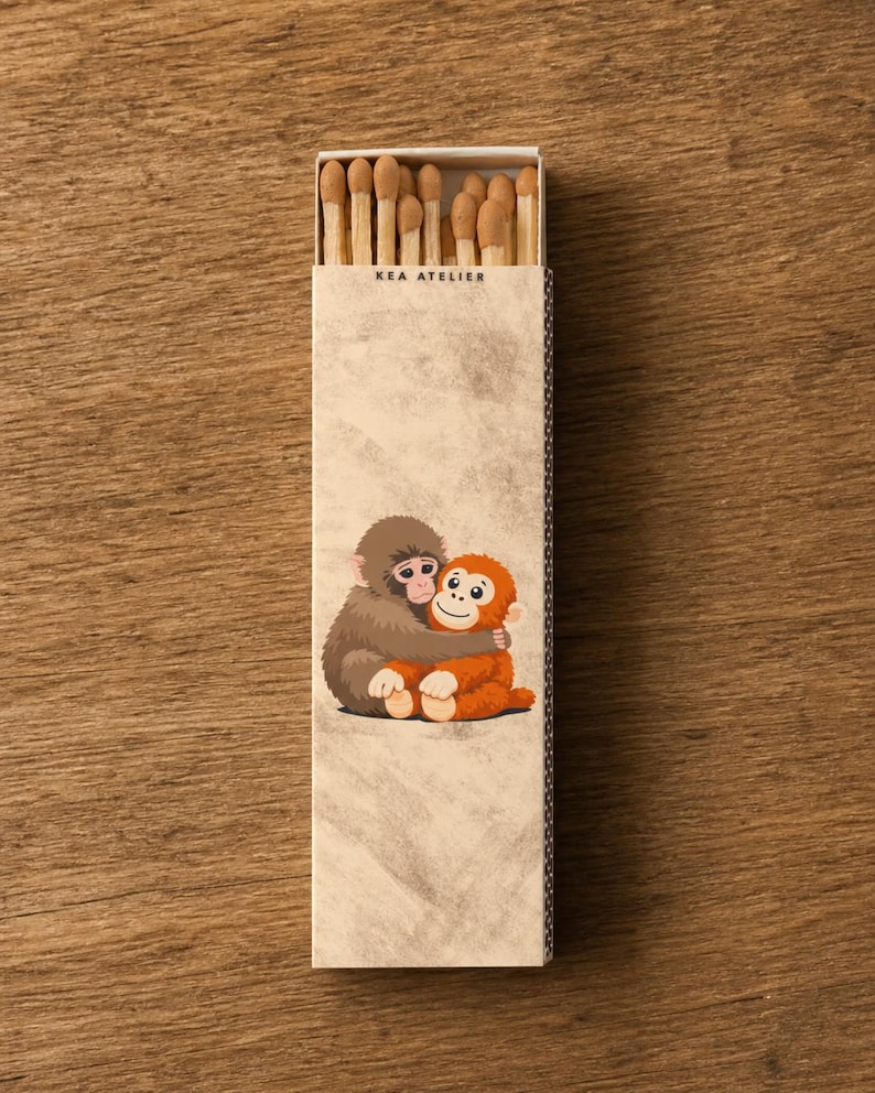 May include: A matchbox with the text "KEA ATELIER" features a cartoon illustration of two hugging monkeys. The match heads are visible at the top. The box is on a wooden surface.
