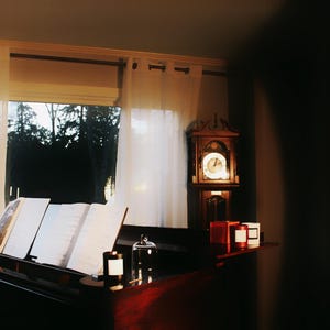 May include: A dimly lit room with a grand piano, sheet music, and a grandfather clock. The piano is open with music sheets. Candles and a glass cloche sit on the piano and a shelf. Sheer curtains frame a window with a view of trees.