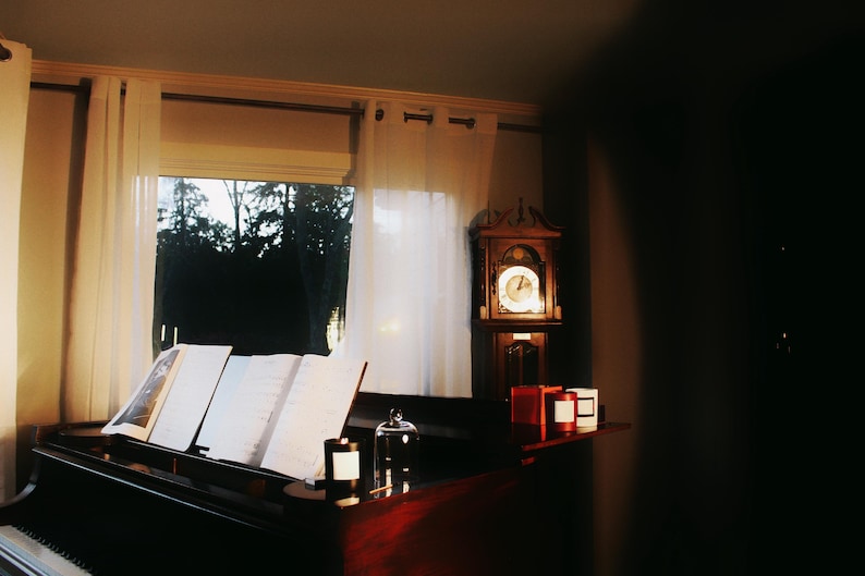 May include: A dimly lit room with a grand piano, sheet music, and a grandfather clock. The piano is open with music sheets. Candles and a glass cloche sit on the piano and a shelf. Sheer curtains frame a window with a view of trees.
